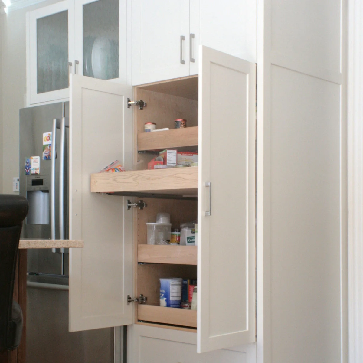 White pantry cupboard doors stand open beside a refrigerator to reveal wood pull-out trays holding food containers and packets.