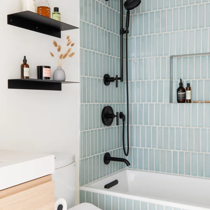 Pale blue vertical tiles and black shower fittings sit beside a white bath, floating black shelves and a light wood vanity edge.
