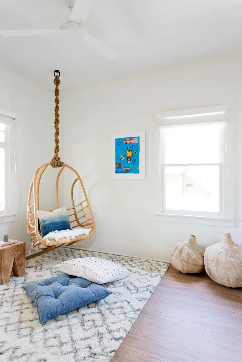 A woven hanging chair with pale and blue cushions occupies a white room corner above a pale patterned rug, beside loose blue floor cushions and woven floor vessels.
