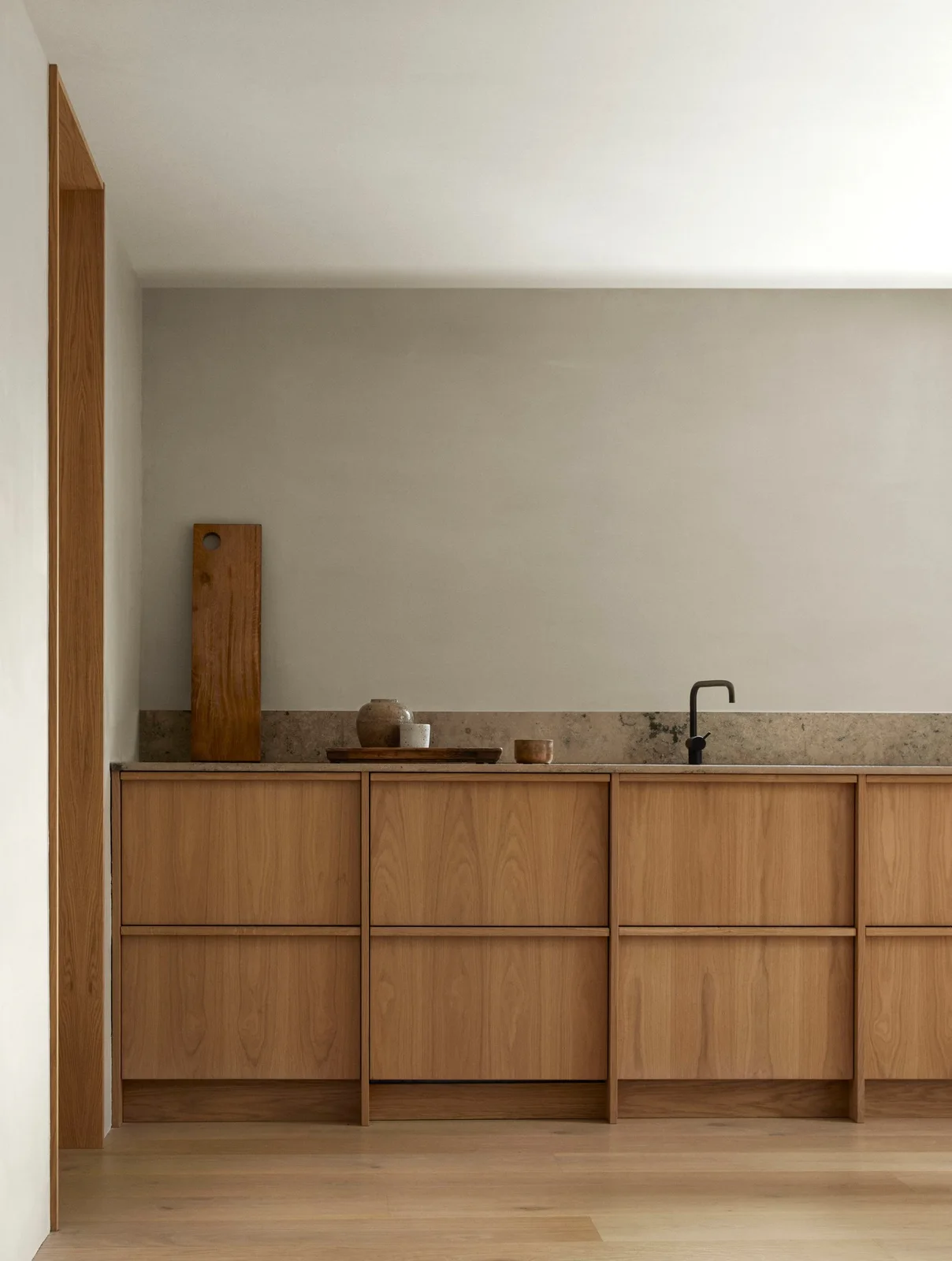 A straight run of wooden cabinets has horizontal rails, a pale mottled counter and backsplash, and a dark tap against a plain pale wall