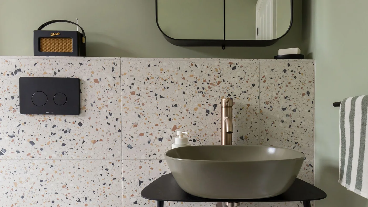 Pale sage walls rise above terrazzo-patterned tiles behind an olive-toned vessel basin, black shelf and mirrored cabinet.