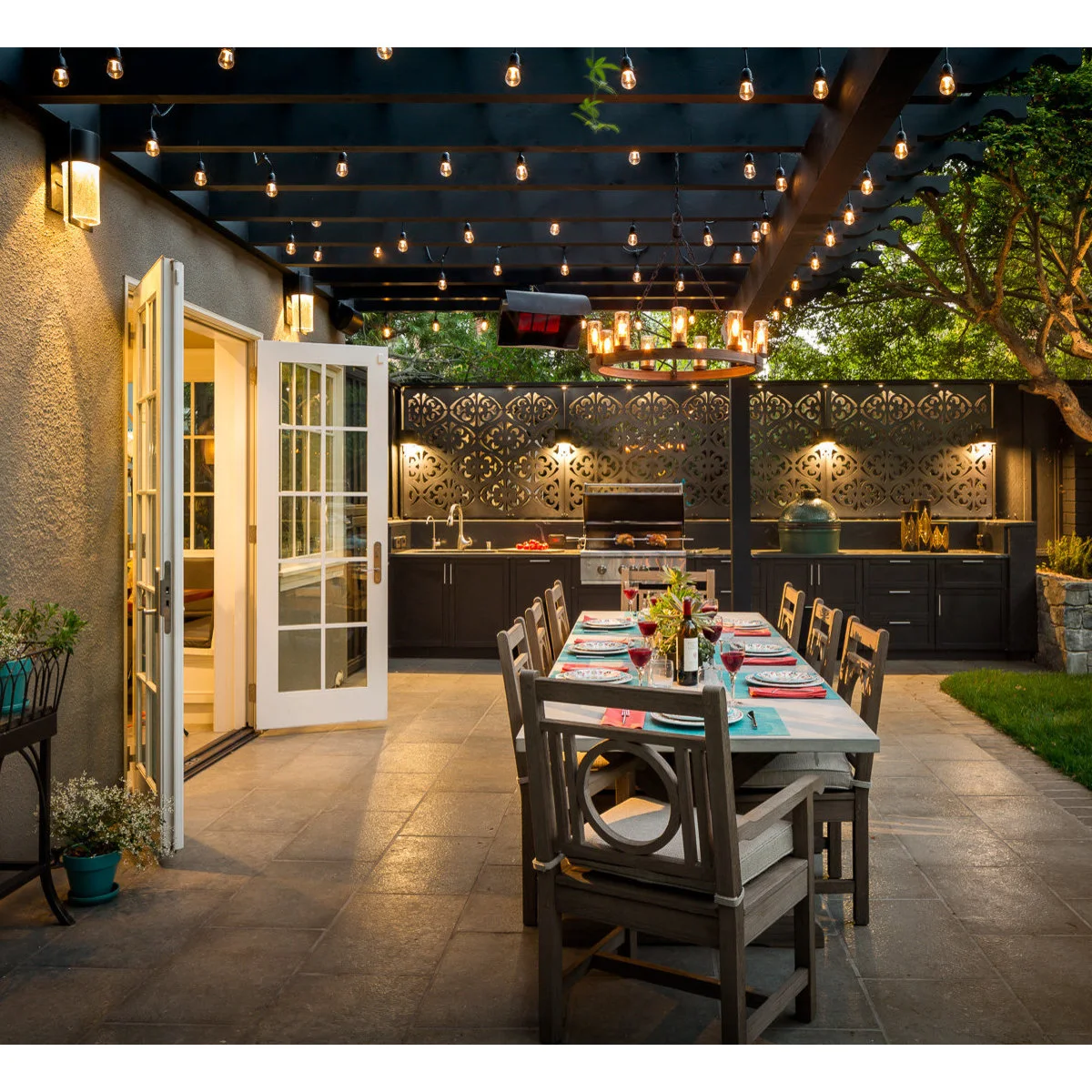 Outdoor dining table beneath string lights facing a dark kitchen run with grill, sink and patterned privacy screen