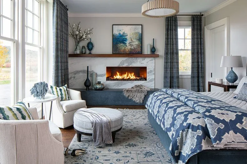A pale veined bedroom fireplace wall with a narrow opening sits below a wood shelf beside two light chairs and blue patterned bedding