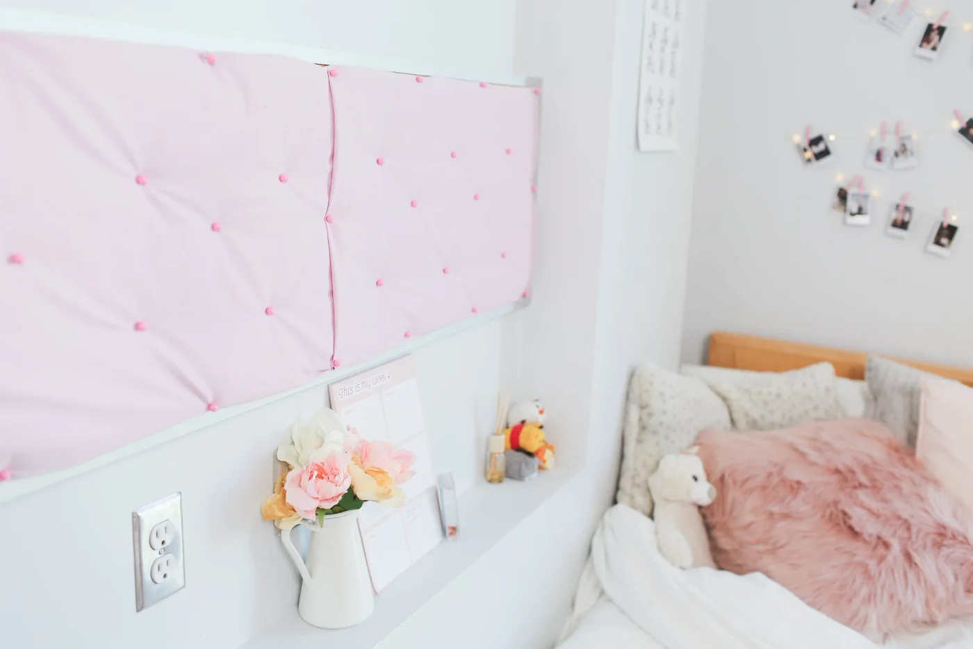 Pink fabric-covered noticeboard above a white ledge beside a dorm bed with pale bedding