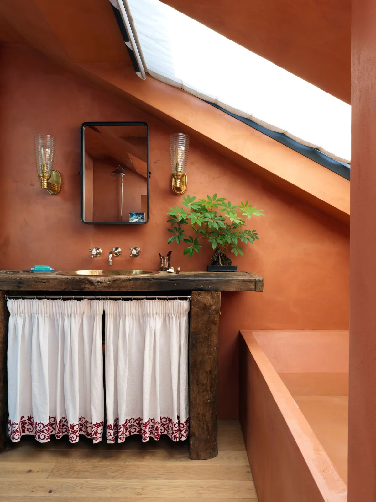 A fabric skirt hangs below a basin beneath rust-colored walls and a sloping window.