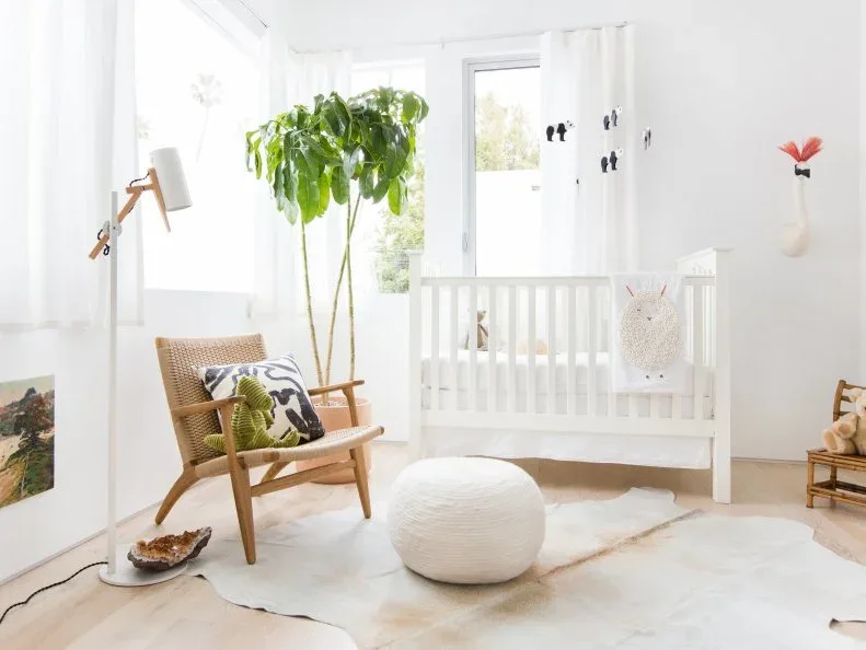 Bright white nursery with white crib, woven wooden chair, pale round pouf, leafy plant and quiet pale floor covering.