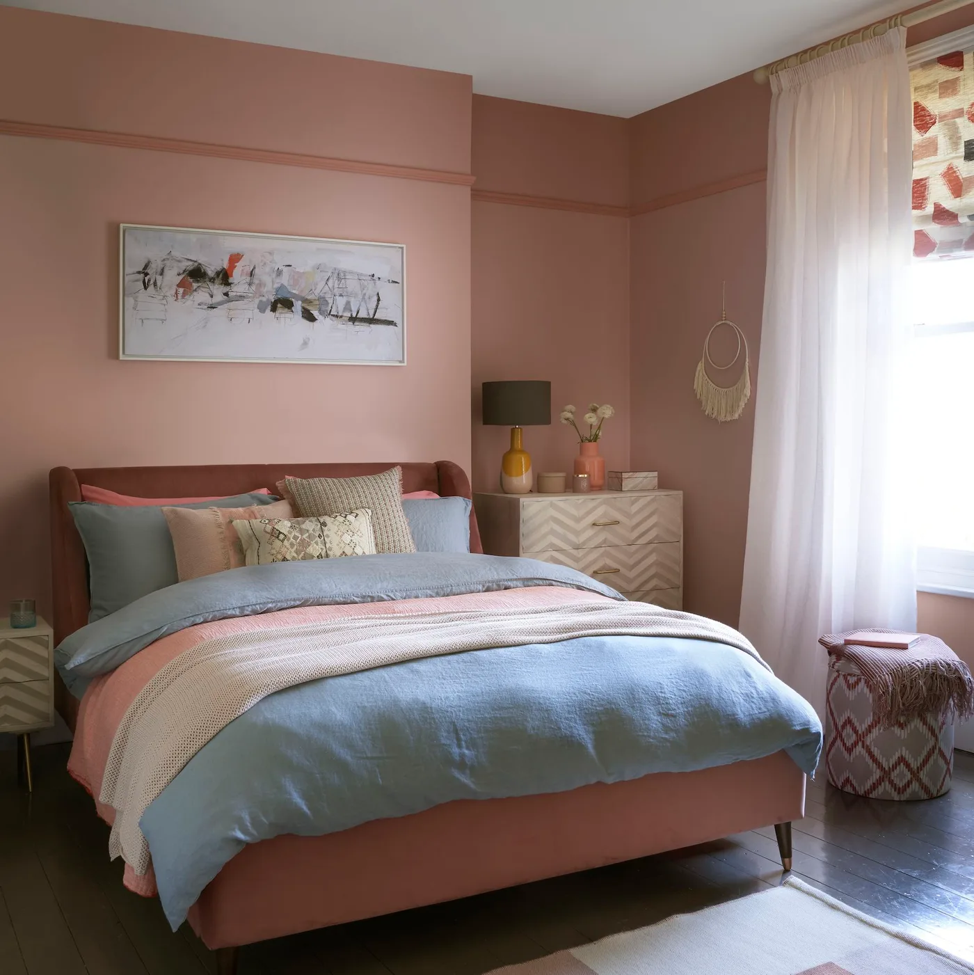 Clay pink bedroom with a straight upholstered bed, blue-gray bedding, a pale geometric drawer chest and a mustard lamp base
