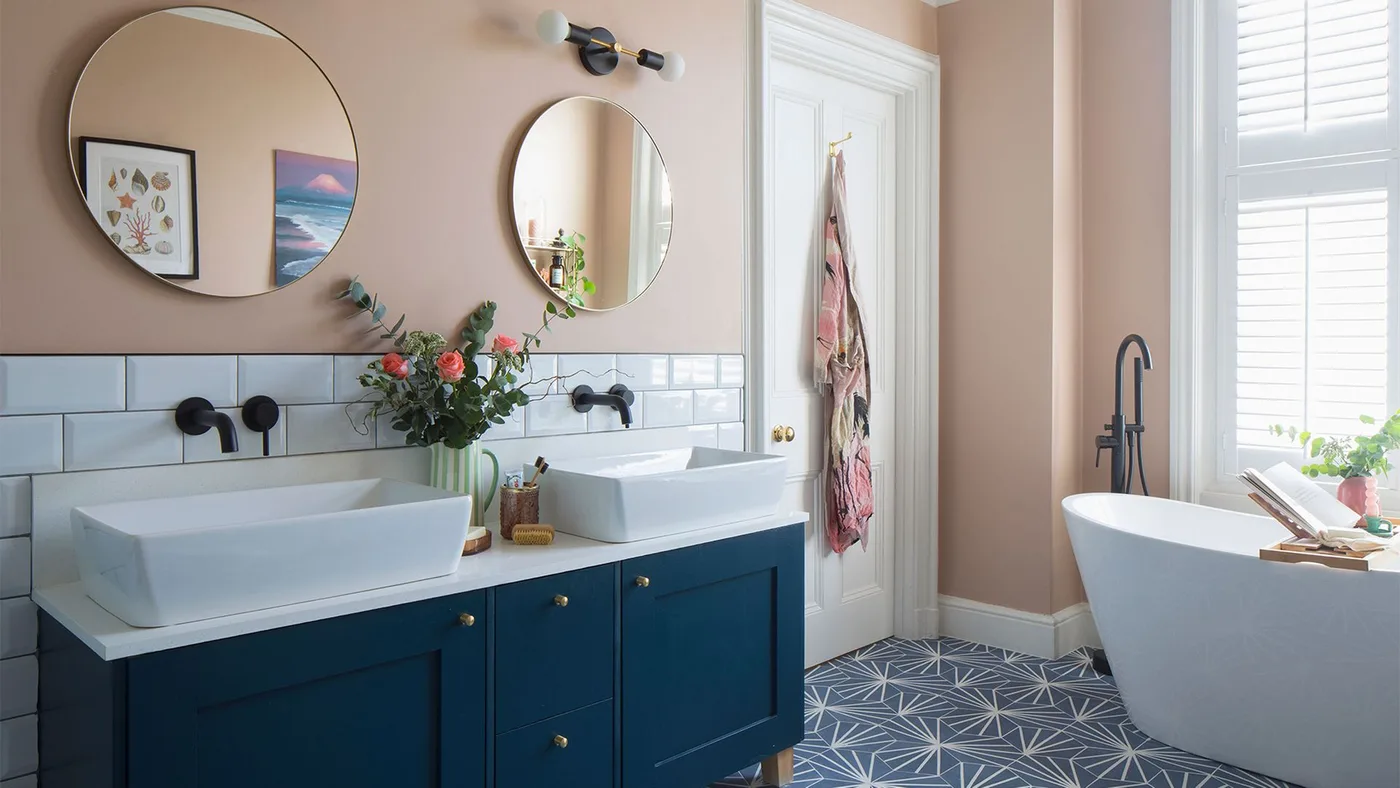 A navy double vanity stands below peach walls and round mirrors beside a white freestanding bath