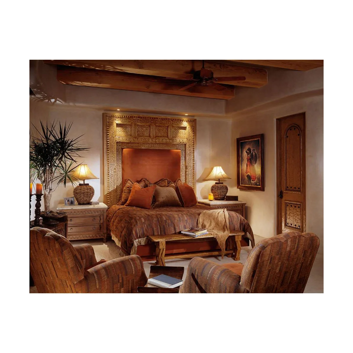 Warm Southwestern bedroom with a large carved headboard surround, orange-brown bedding, ceiling beams and two upholstered chairs