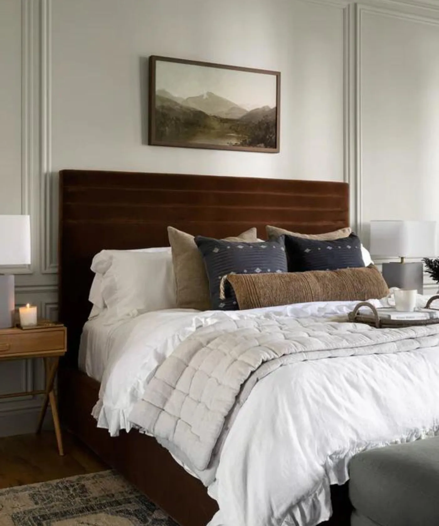 Bedroom with dark brown upholstered headboard, white bedding, pale checked blanket, dark blue cushions and warm bedside tables.