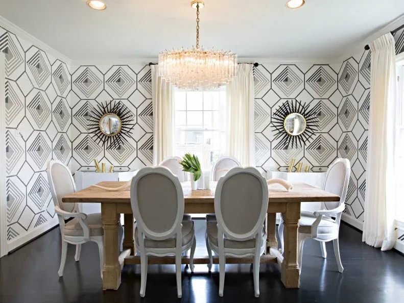 Dining room with outlined geometric wallpaper, pale oval-backed chairs, a long wood table, twin sunburst mirrors and white curtains