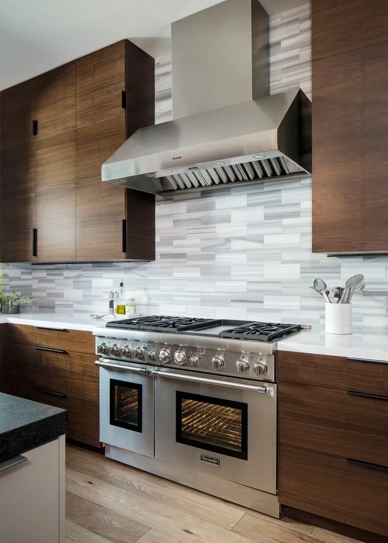 A stainless range and broad hood sit between walnut cabinets with black pulls and gray-and-white rectangular backsplash tiles.