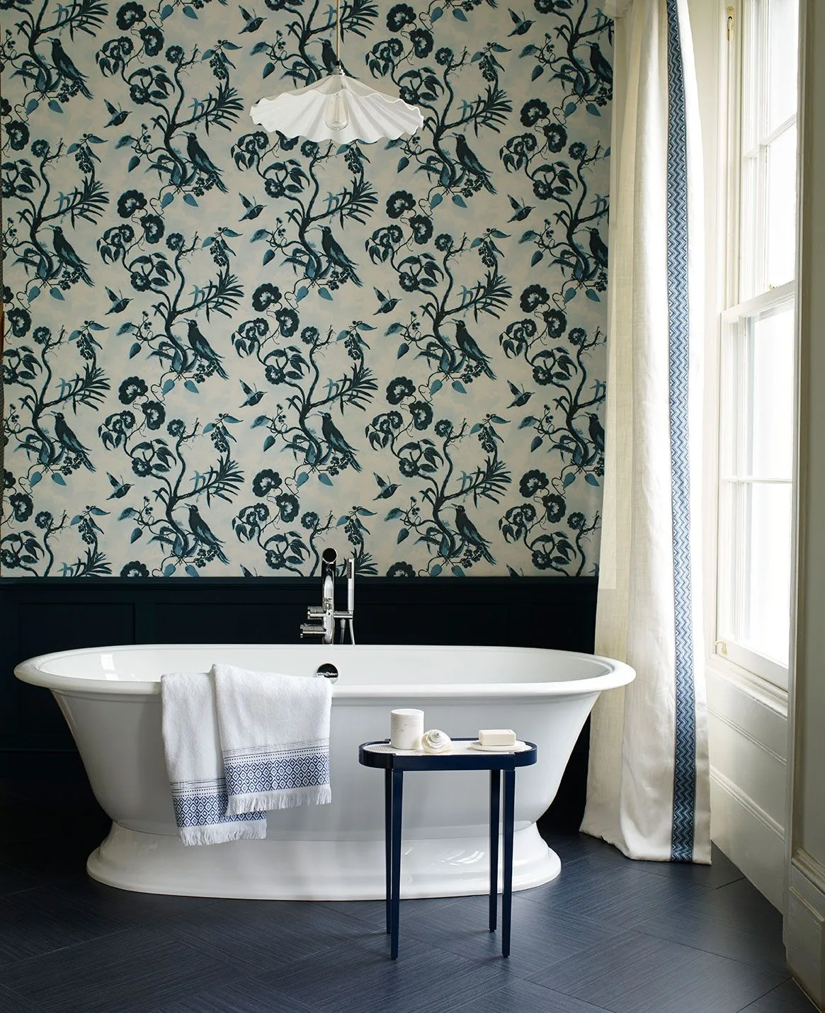 Bathroom with a blue floral wall pattern, dark lower paneling, white freestanding bath, pale curtain and a small blue side table.