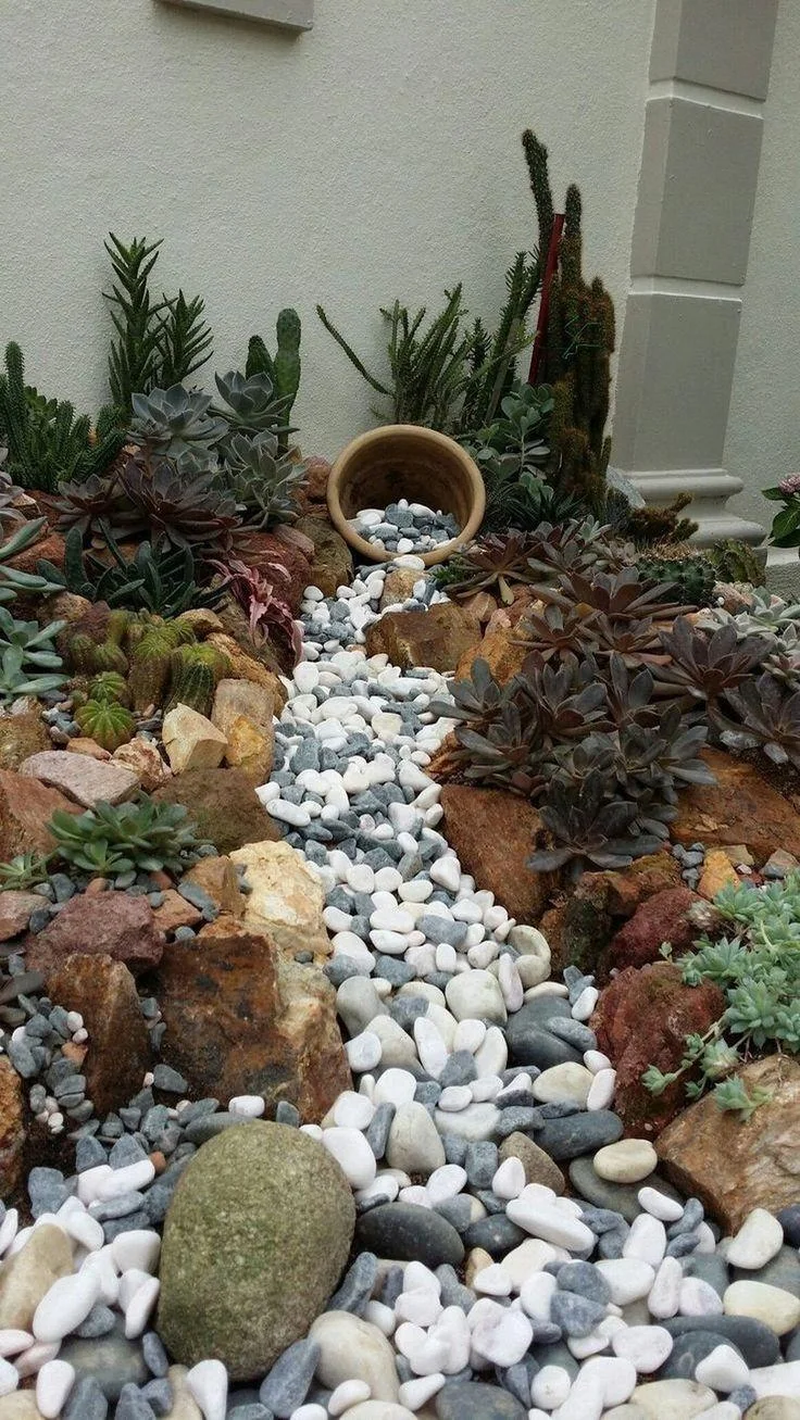 A tipped terracotta pot appears to spill white and gray pebbles through a succulent bed