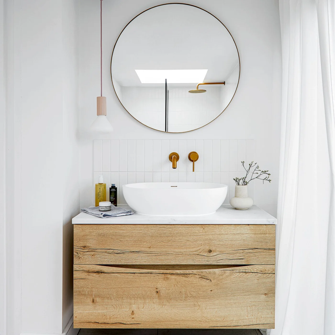 A small wood-fronted vanity with a white vessel basin sits below a round mirror beside gold-toned taps and a slender pendant