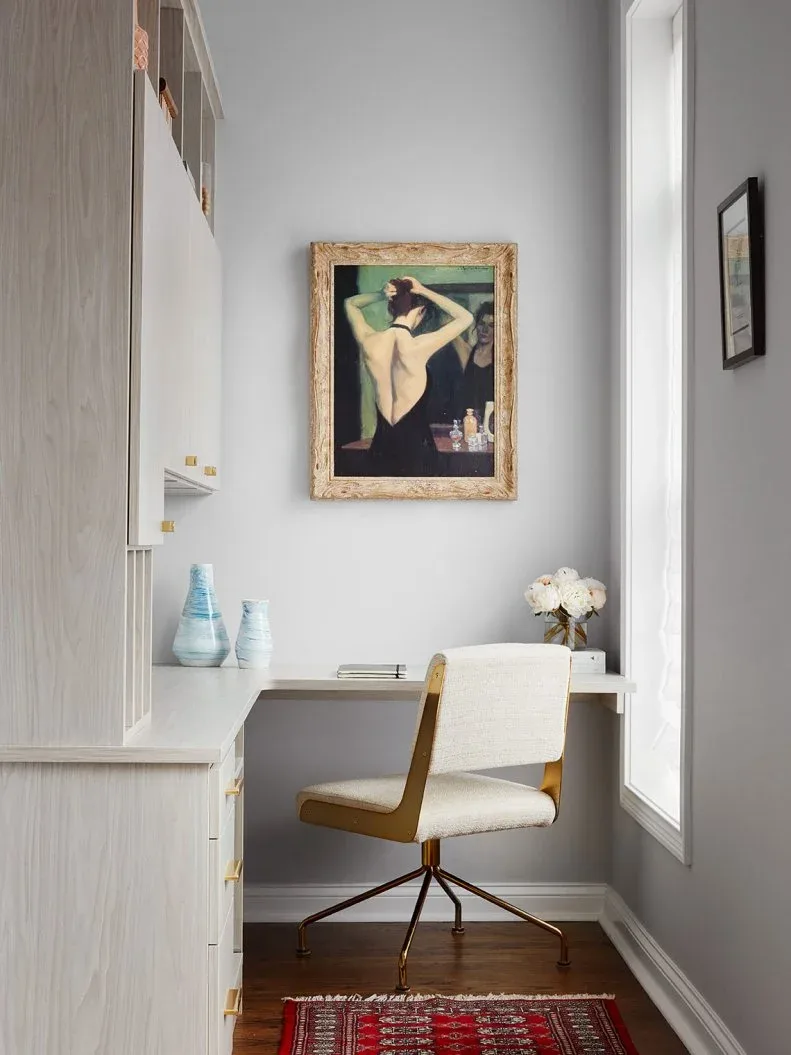 A pale fitted desk occupies a narrow recess beside tall cabinetry and a window, with a swivel chair and framed portrait.