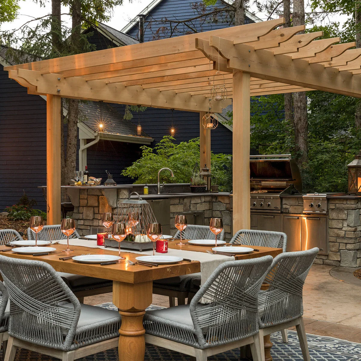 Outdoor dining table and woven chairs beside a pergola-covered kitchen with stone-faced bases and metal grill