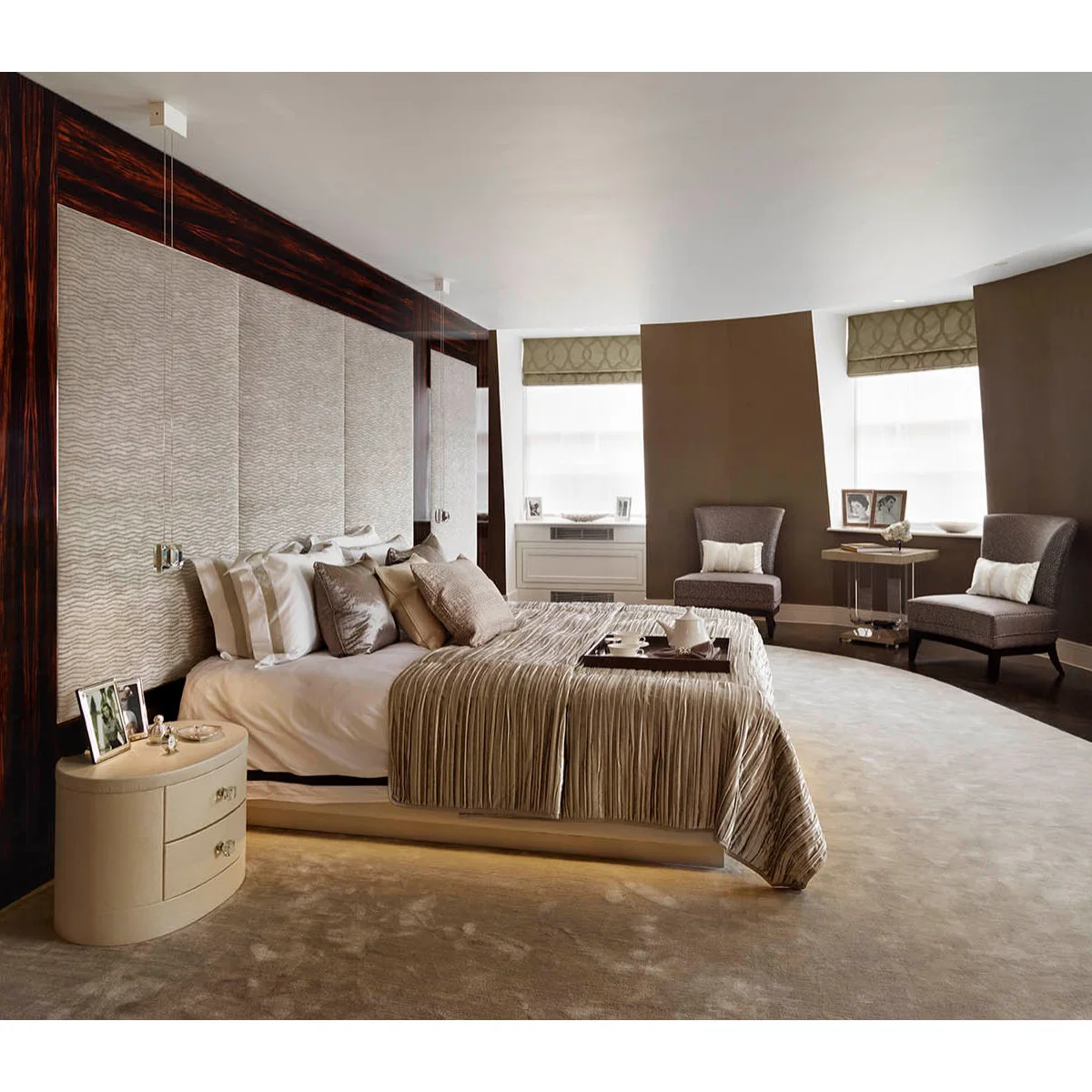 Brown bedroom walls with a broad pale headboard, striped brown bedding and two chairs near the windows.