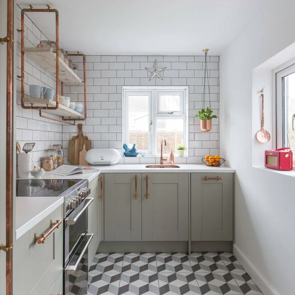Light gray lower cabinets with copper-toned pulls sit beneath white rectangular tiles beside glazed wall cupboards and a geometric patterned floor