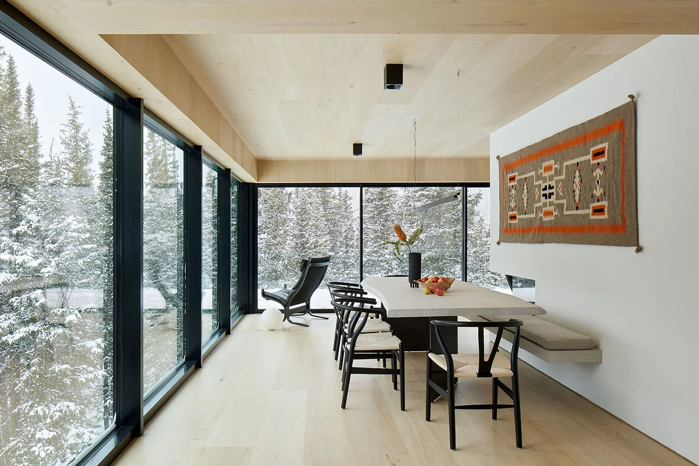 Dark wooden chairs surround a pale table beside a cushioned wall bench, large windows, wooden ceiling panels and a woven wall hanging