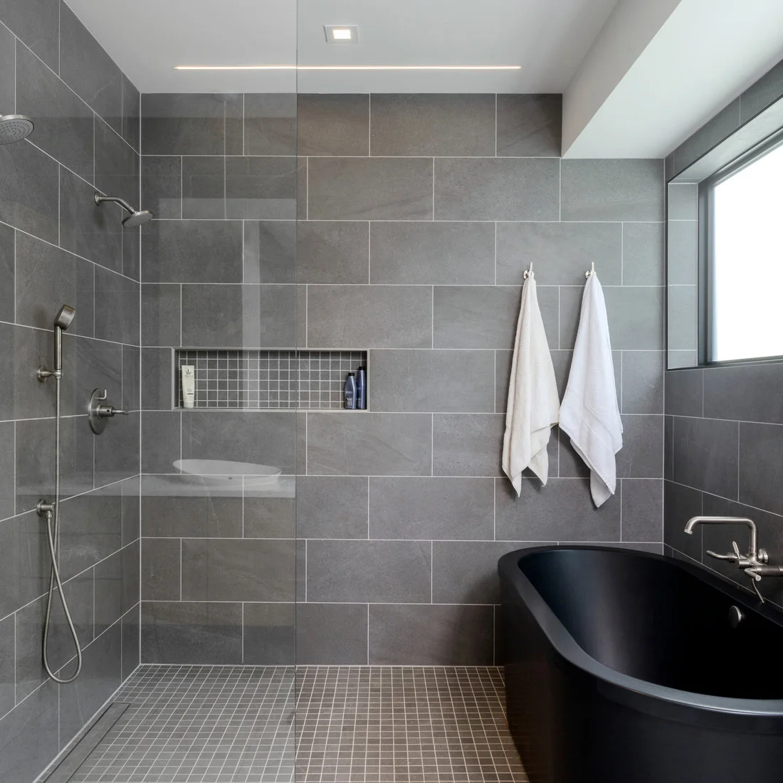 Small gray square floor mosaics meet broad gray wall tiles and a black freestanding tub.