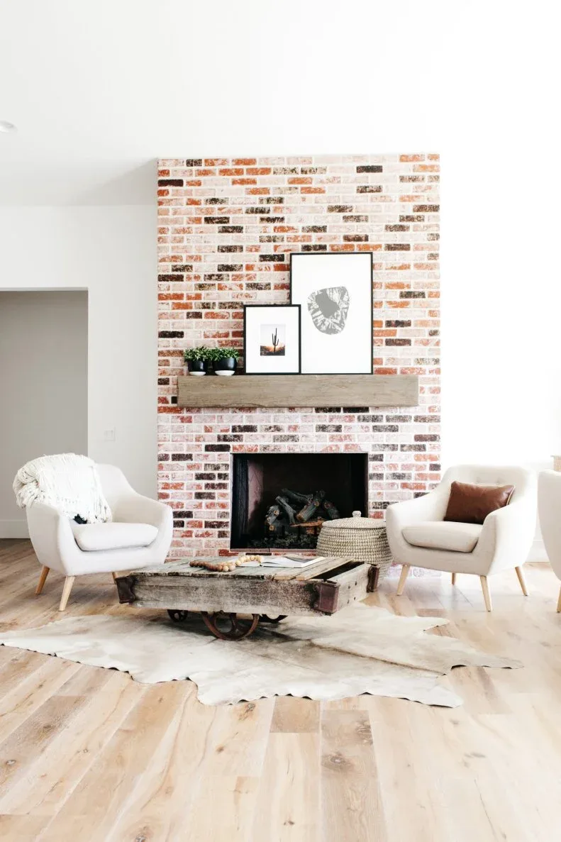 Red-and-white brick fireplace with a broad wooden mantel between two pale armchairs.