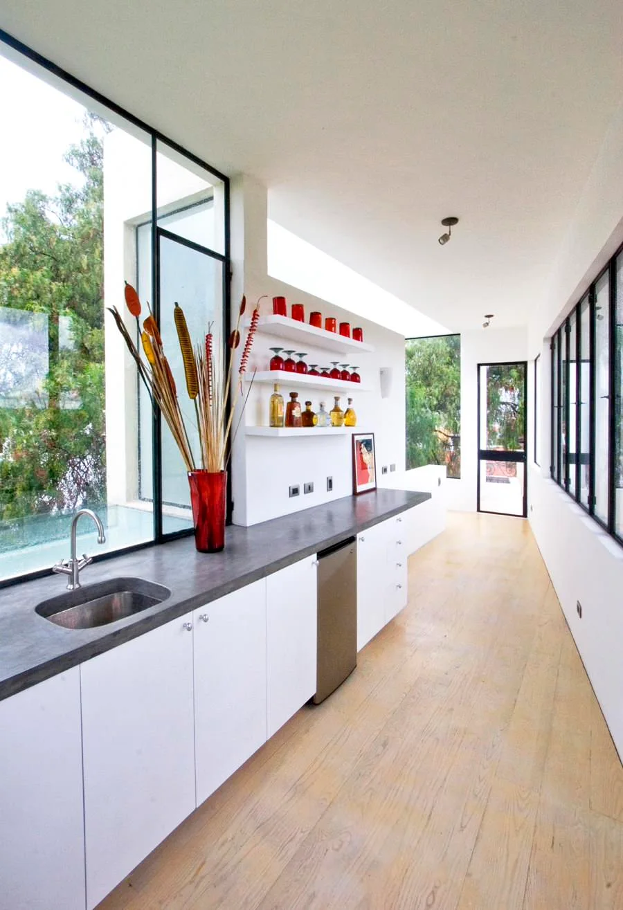 Red glasses on white shelves above a dark counter in a narrow white kitchen with black-framed windows and a red vase.
