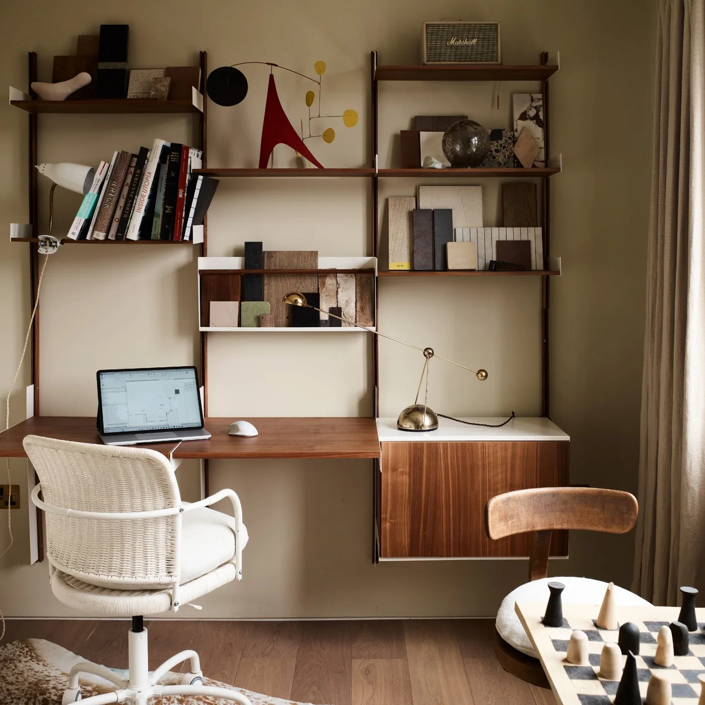 A warm beige room has a wood wall-mounted shelf and desk system, a closed cabinet, a pale woven swivel chair and books and samples on the upper shelves.