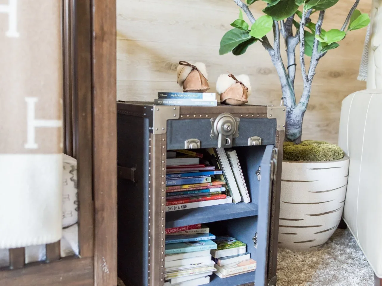 An open trunk-style side cabinet holds children’s books beside a nursery chair and a leafy plant.
