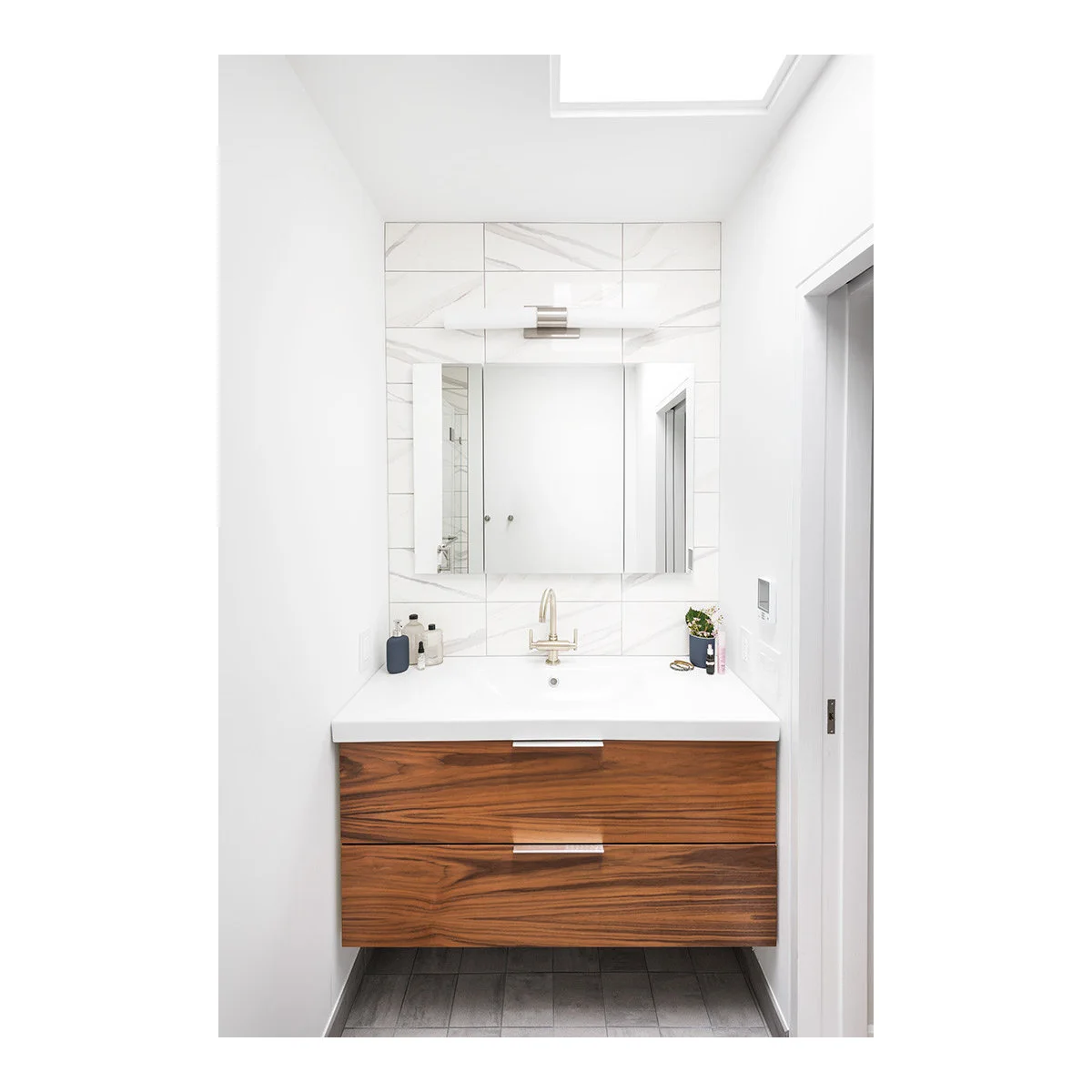 Wood-fronted floating vanity fitted between white walls below a mirrored cabinet and veined tile backsplash