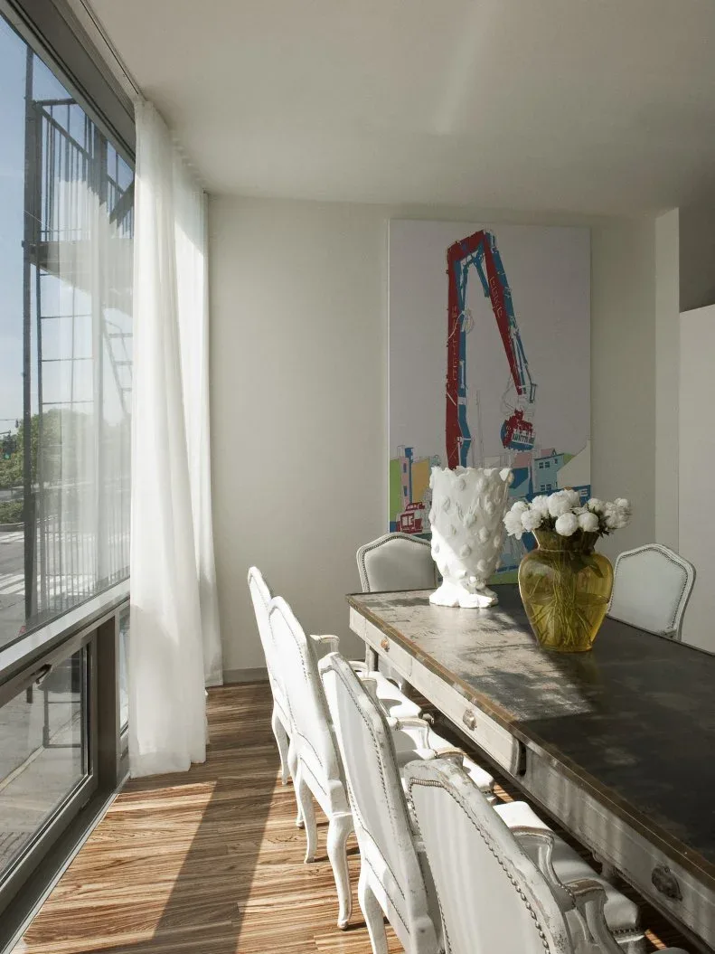 Long dark dining table with pale upholstered chairs beside tall glazed windows and sheer curtains