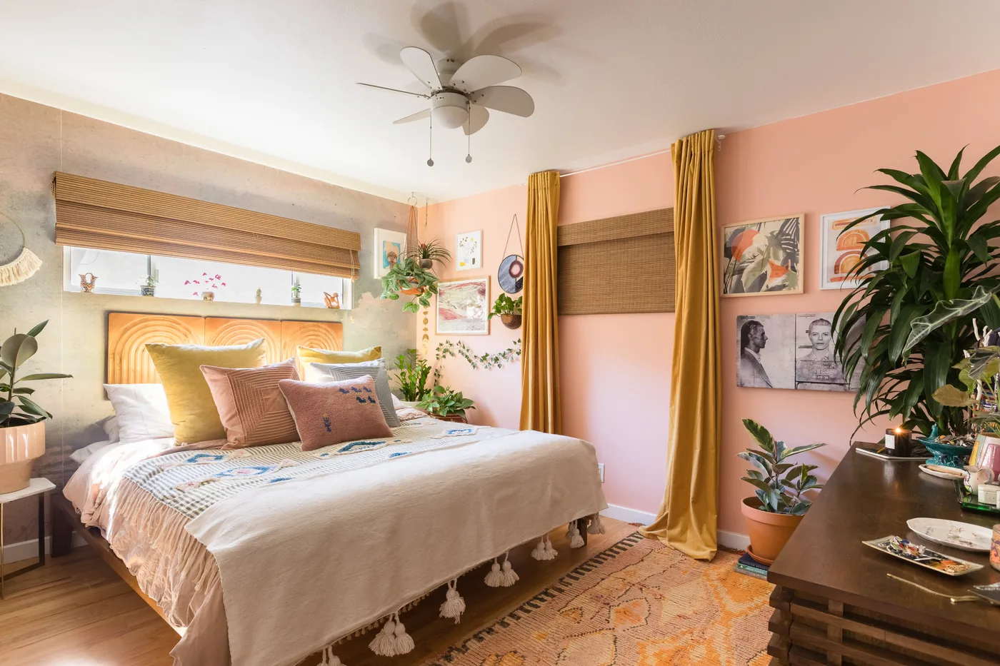 A pale bed stands beside pink walls, mustard curtains, woven window shades and a dark wood dresser with leafy plants and framed pictures nearby