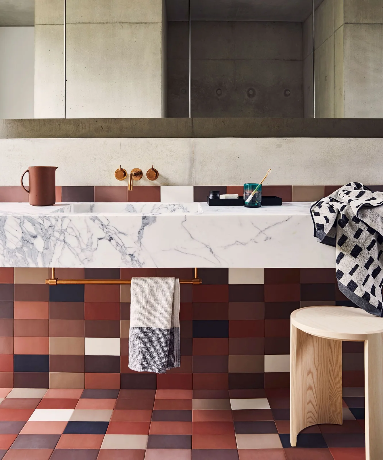 A broad pale veined counter sits above multicolored square wall and floor tiles beside a large mirror, warm-toned fittings and a small wood stool