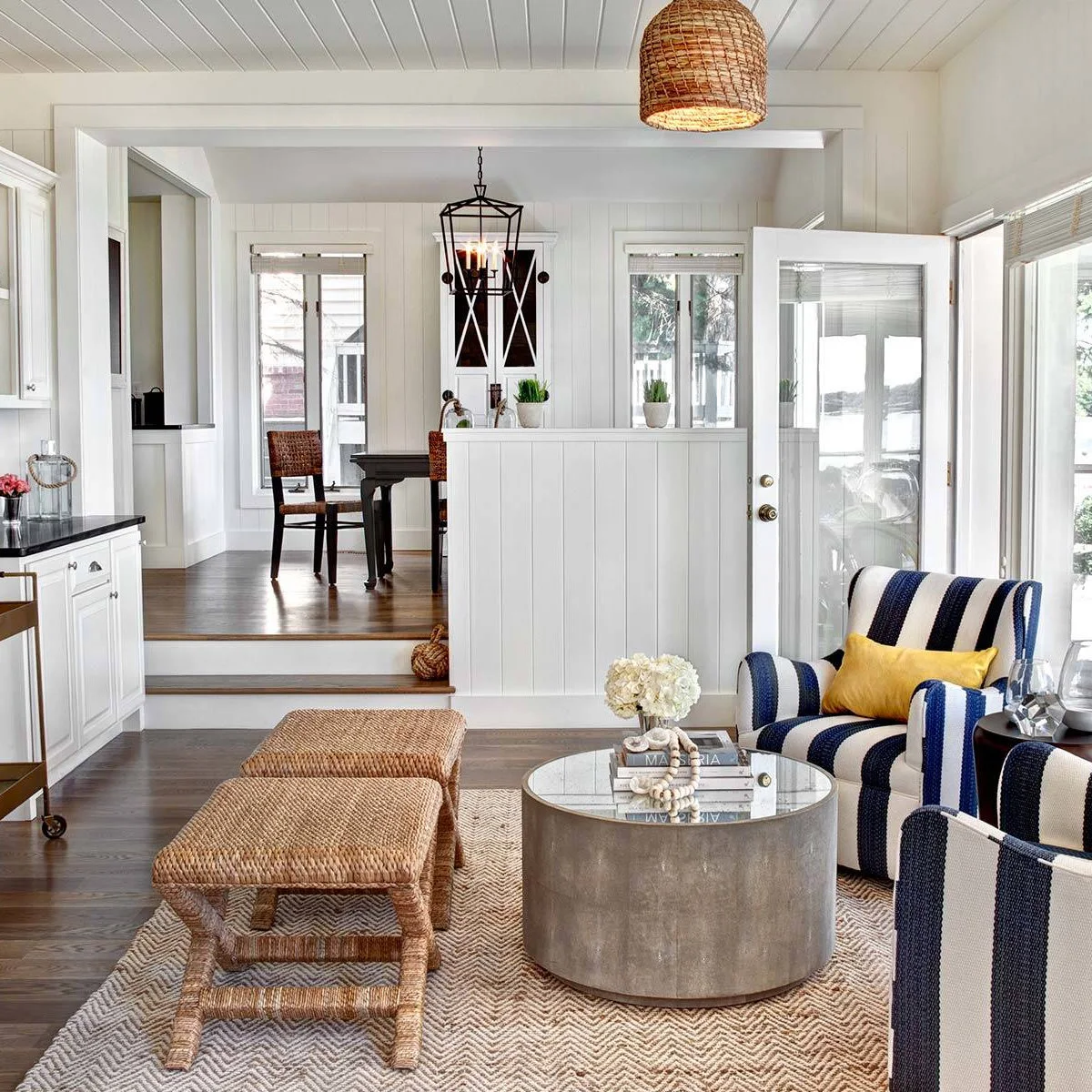 White paneled living room with striped armchairs, woven stools and a round metal table