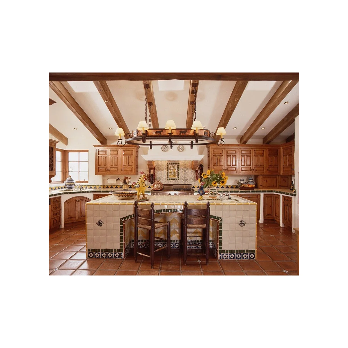 Cream tiled kitchen island with colorful border tiles, wood stools and a patterned cooking wall beneath wood cabinets and beams.