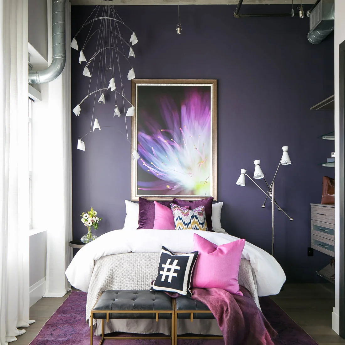 A white bed with purple and pink textiles sits against a dark purple wall beneath exposed silver ductwork and a colorful framed picture