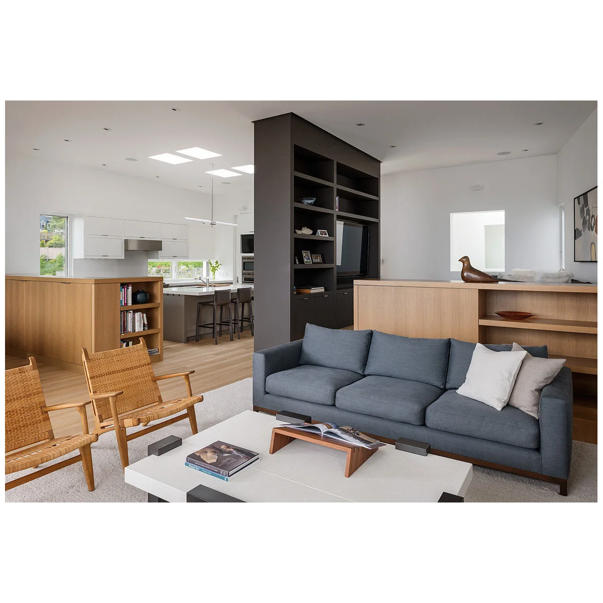 A dark tall shelving unit and lower wood cabinetry divide a white kitchen from a living room with a blue sofa.