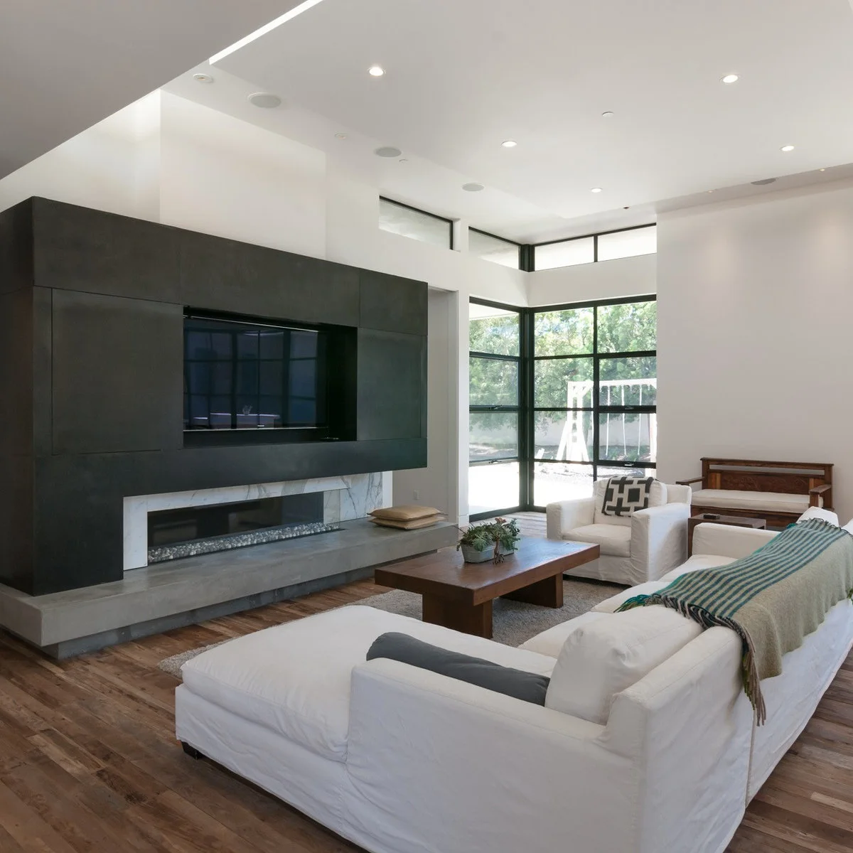 A recessed television in a large dark wall panel above a narrow fireplace, with white seating and tall black-framed windows.