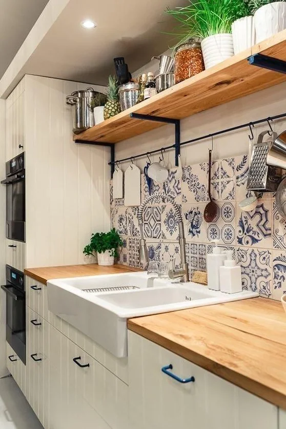 Pale paneled cabinets and a wood-toned counter sit below blue patterned backsplash tiles with a white sink, utensil rail and long upper shelf