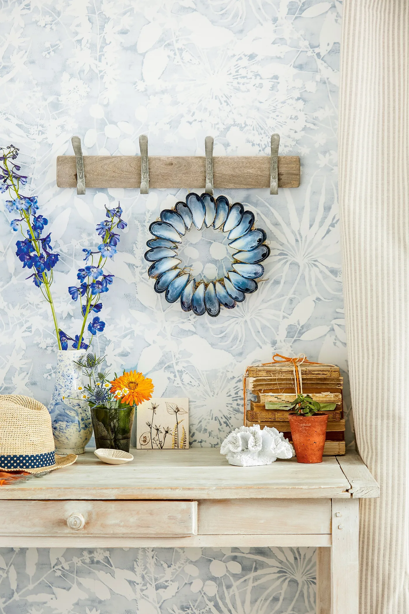 Console corner with pale leafy wall pattern, wooden drawer table, simple wooden peg rail, flowers and a decorative blue-edged mirror.