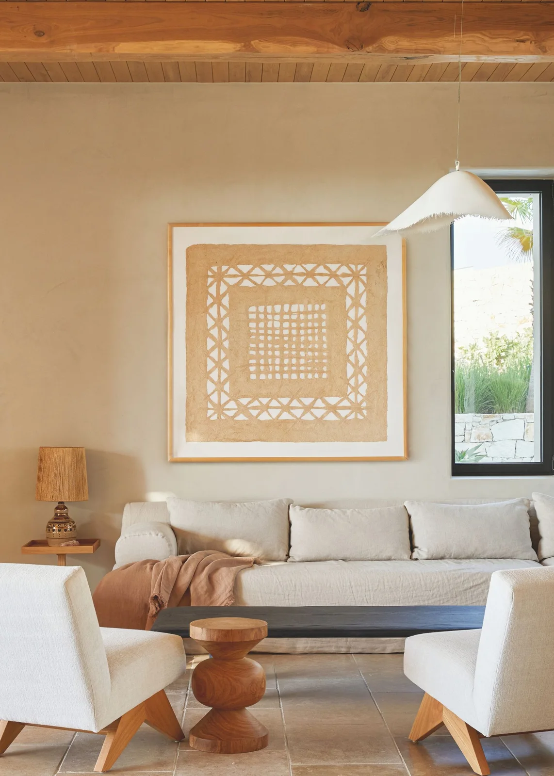 Pale textured living-room seating surrounds a low dark table beneath wood ceiling details beside graphic artwork, a shaped wooden stool and pale pendant