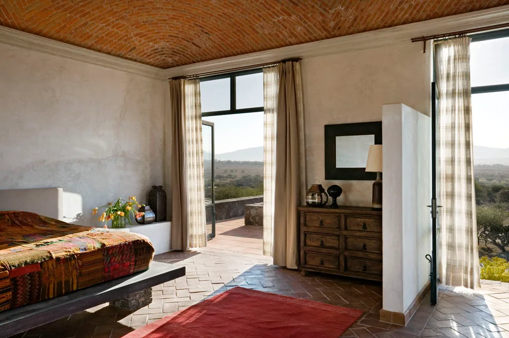 Colorful patterned bedcover on a low bed beside pale walls, sheer curtains and a wood chest under a brick ceiling.
