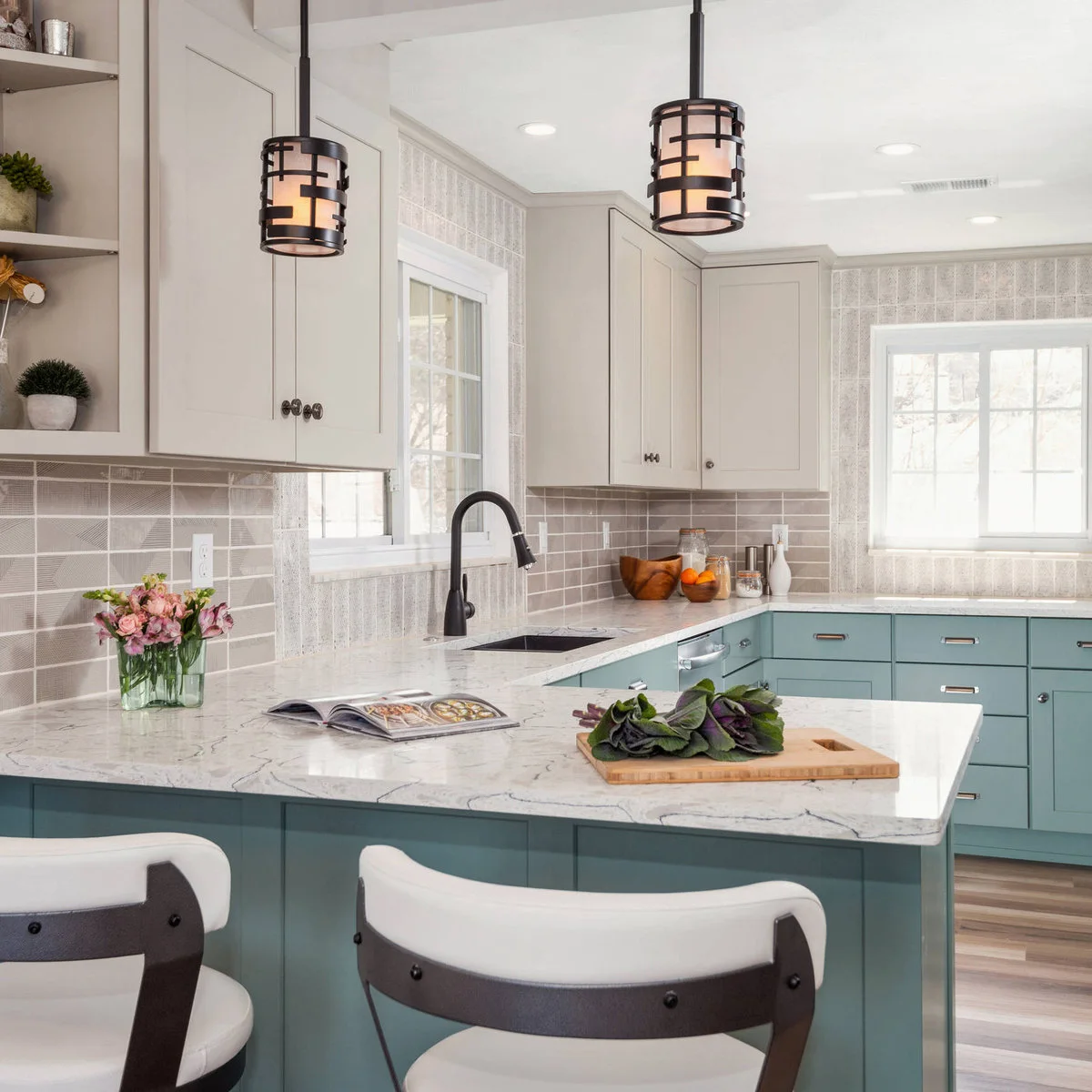 Muted turquoise lower cabinets and a peninsula sit beneath creamy pale cupboards beside gray tile and dark pendant frames.