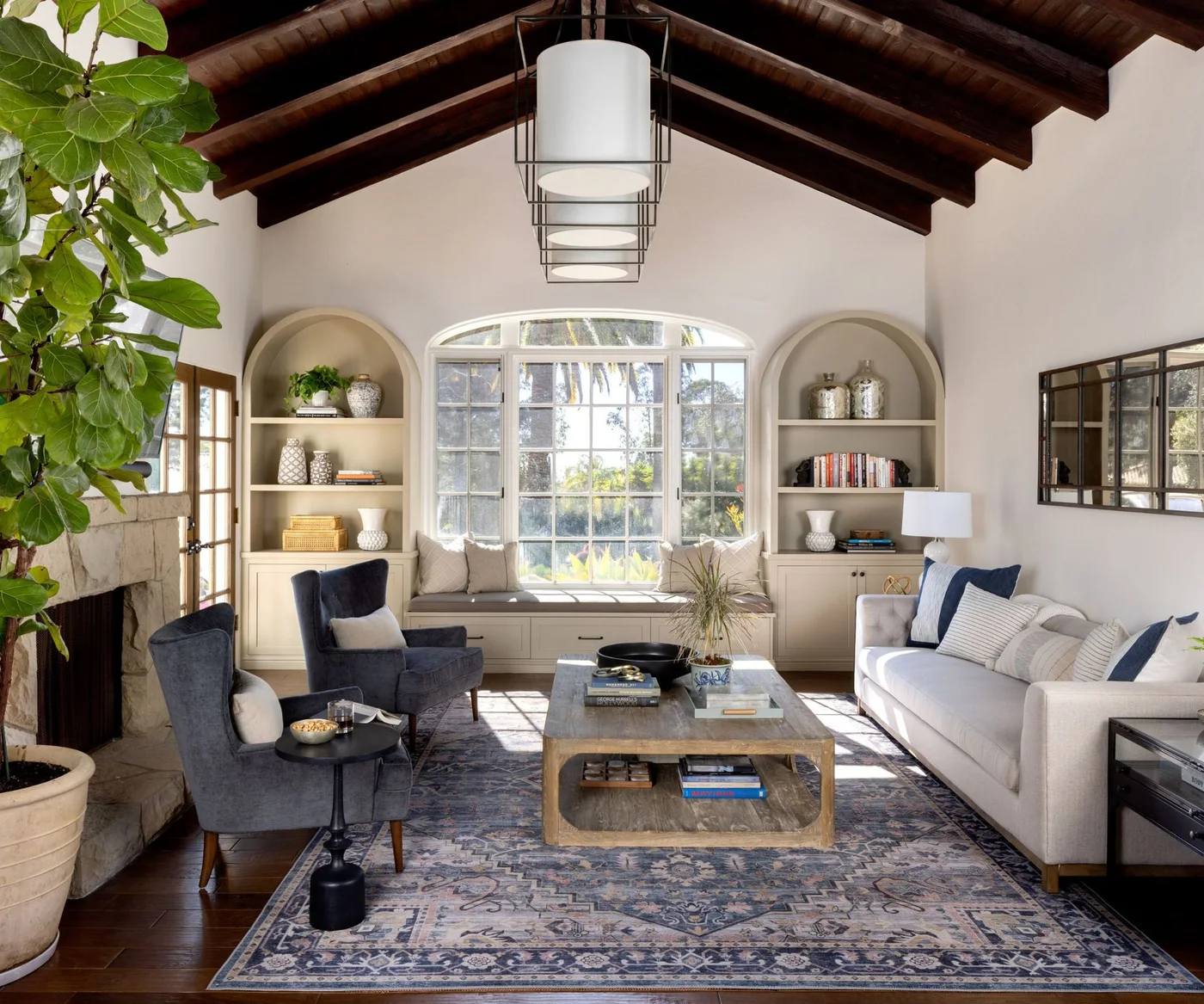 Arched living-room shelves flank a window seat beneath dark wooden ceiling beams beside blue-gray chairs, a pale sofa and patterned rug