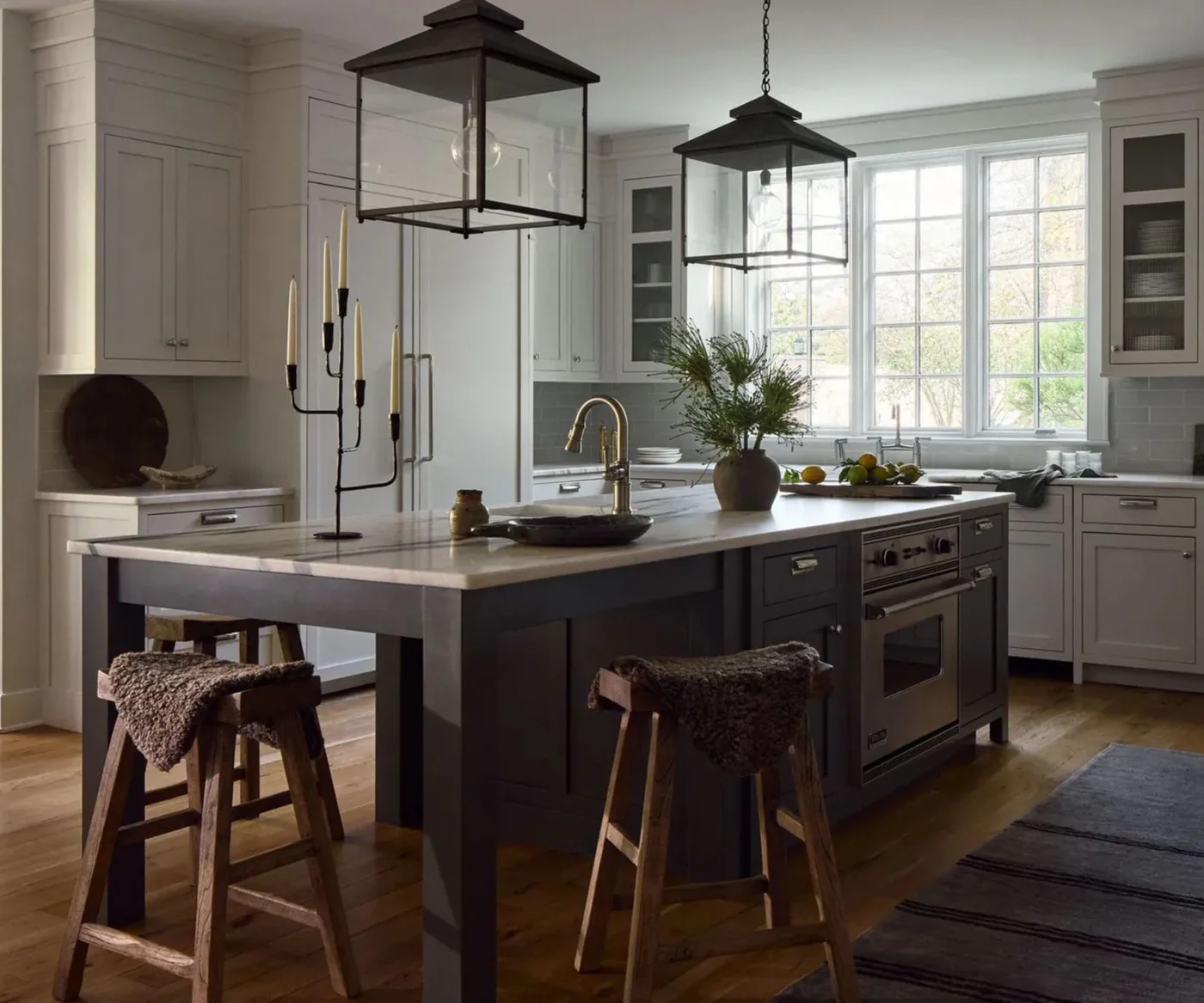 A white framed kitchen with a broad window has a dark island containing an oven, two large black lanterns, wood stools and a pale tiled backsplash.
