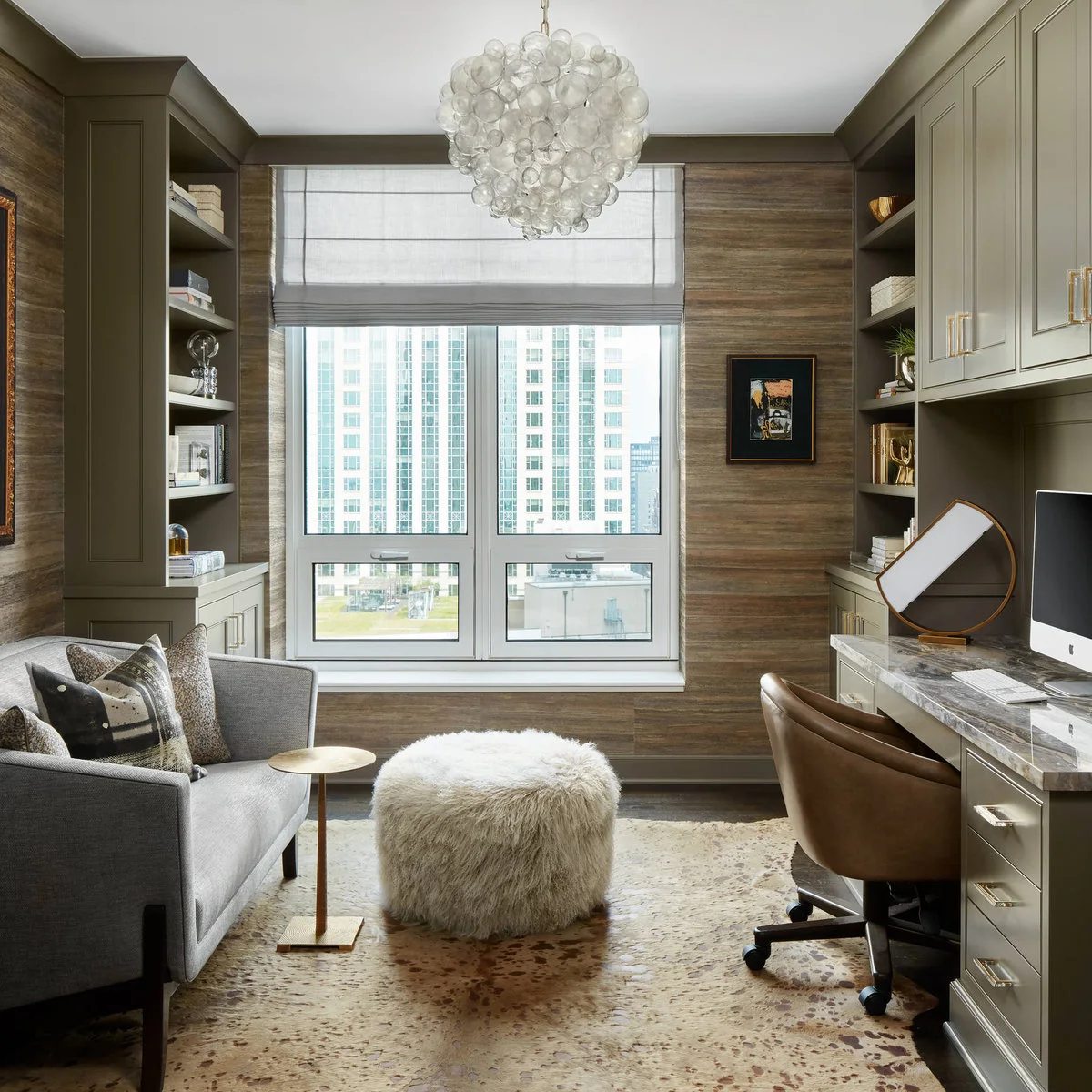 A gray loveseat and round ottoman opposite a fitted computer desk with upper cupboards and side shelves.