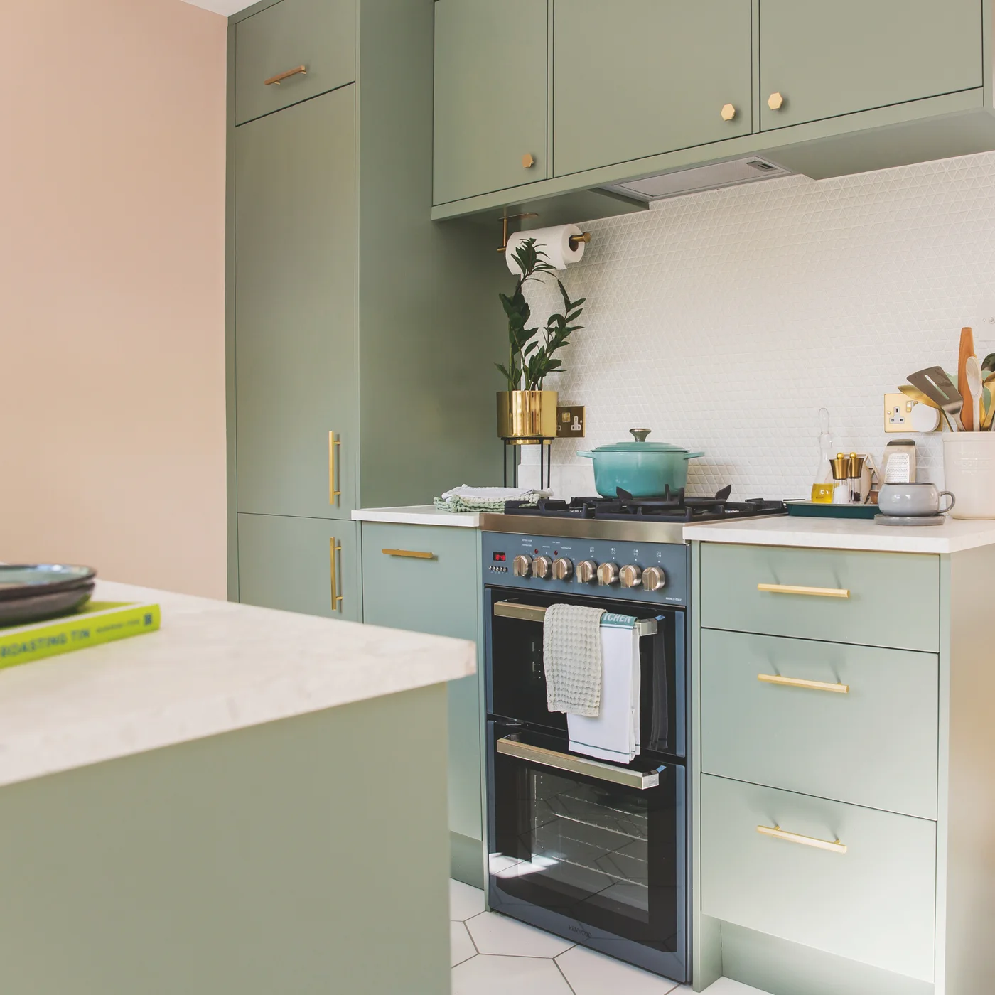 Sage green flat-fronted cabinets with warm metal handles surround an oven and cooking surface beside a pale backsplash