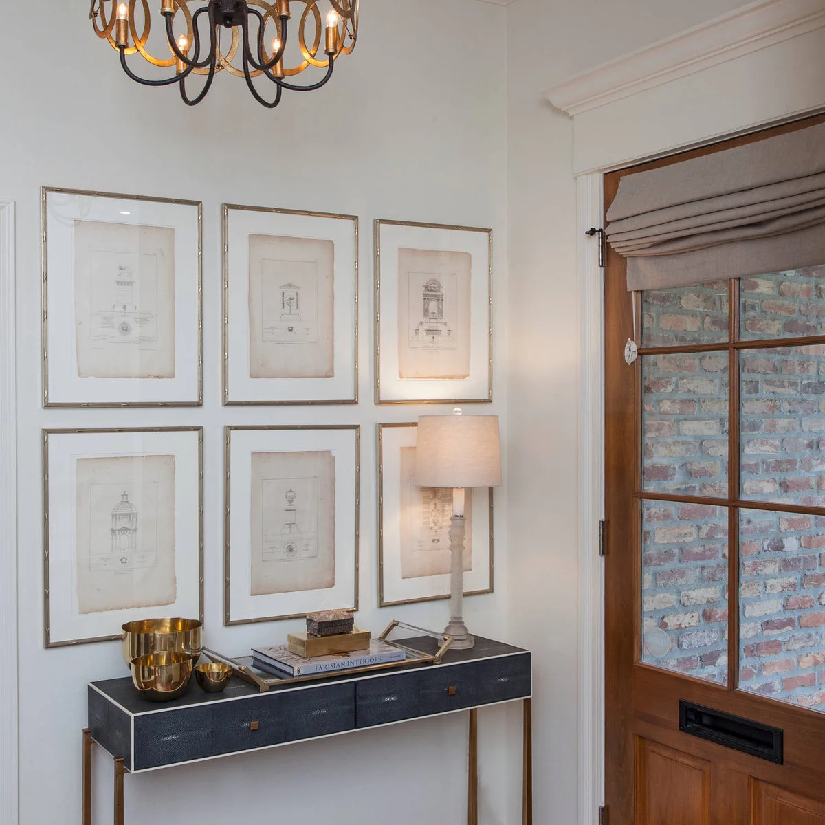 A dark narrow console beneath six evenly arranged pale prints, with a slim lamp and nested warm-metal bowls.