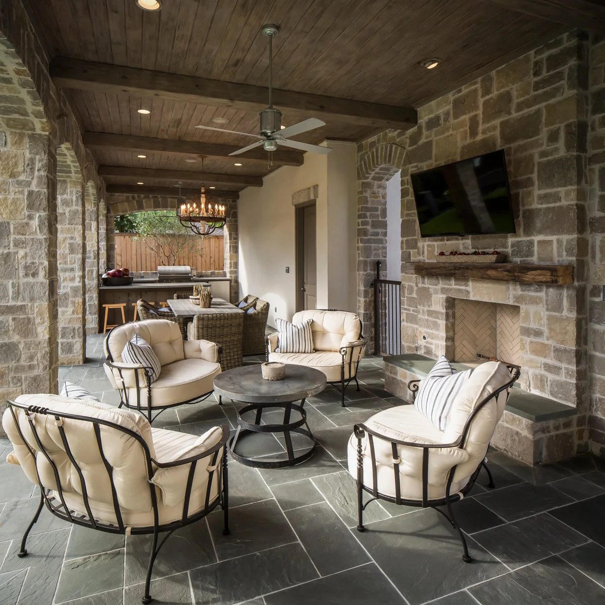 Four pale cushioned chairs surround a round table beneath a wood ceiling, with dining farther along the stone-walled patio.