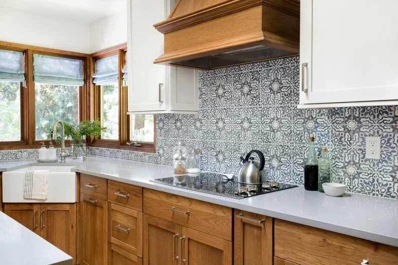 Wood lower cabinets and a matching hood sit beneath white uppers, with a blue-and-white patterned backsplash and corner sink window.